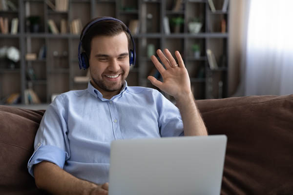 Meeting online. Happy loving young husband wear headphones talk chat to wife say hi using conference video call app on laptop. Smiling man relax on sofa wave hand hello greet friend using webcam on pc