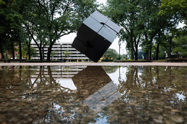 The Cube and surrounding trees are reflected in a puddle on the pavement in Regents Plaza after a rainy morning on campus.